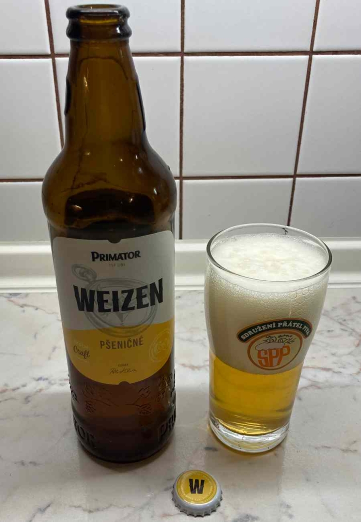 Nalité pivo Primátor Weizen ve sklenici vedle láhve s hustou pěnou